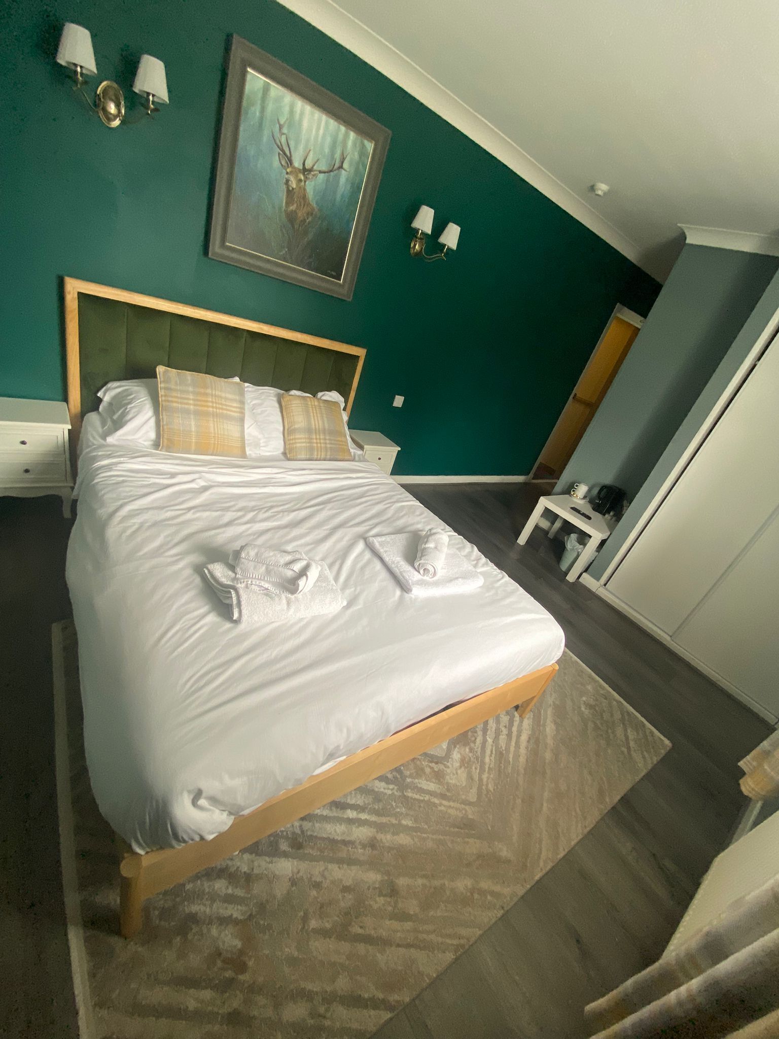 A neatly made bed with white linens and towels, a green upholstered headboard, and a framed deer painting on the teal wall.