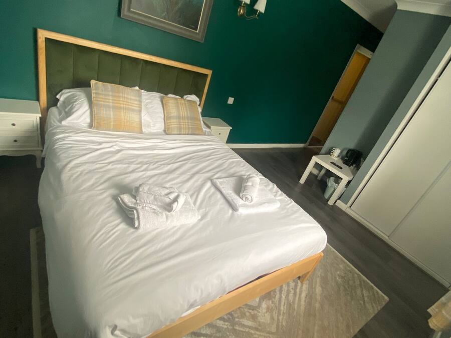 A neatly made bed with white linens and towels, a green upholstered headboard, and a framed deer painting on the teal wall.