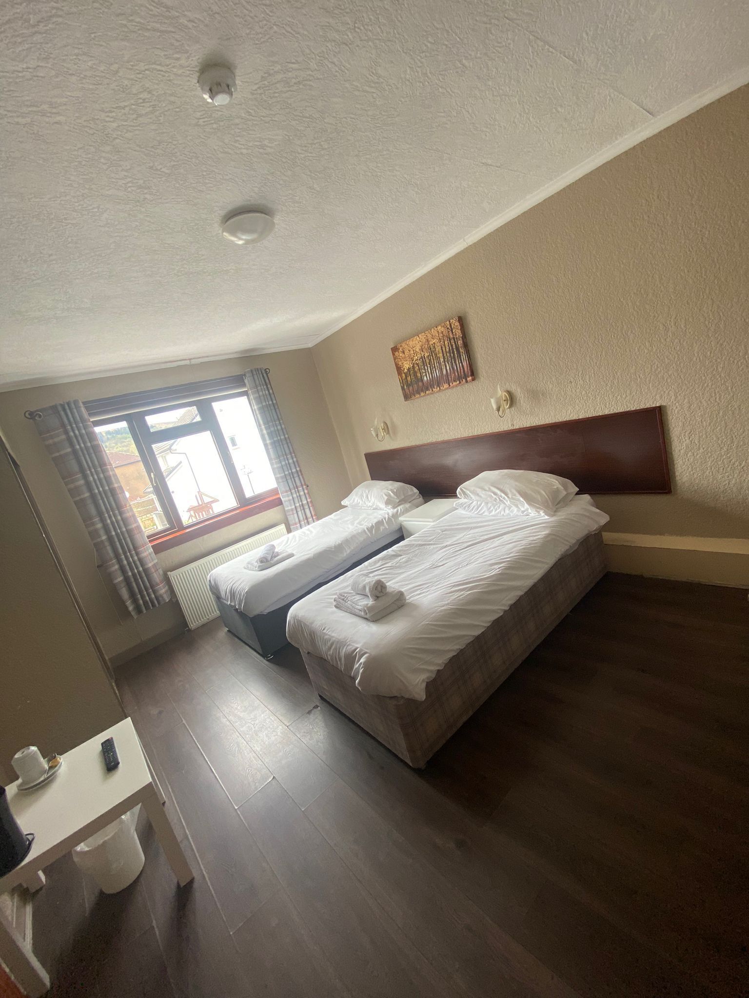 Ein Hotelzimmer mit zwei Betten, einem Fenster mit Vorhängen, einem Heizkörper, einem kleinen Tisch mit Tasse und Fernbedienung sowie einem Wandbild.