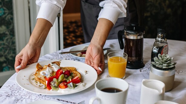 Una persona sirviendo una comida con un plato de alimentos, una cafetera, un vaso de jugo de naranja y un periódico sobre una mesa.