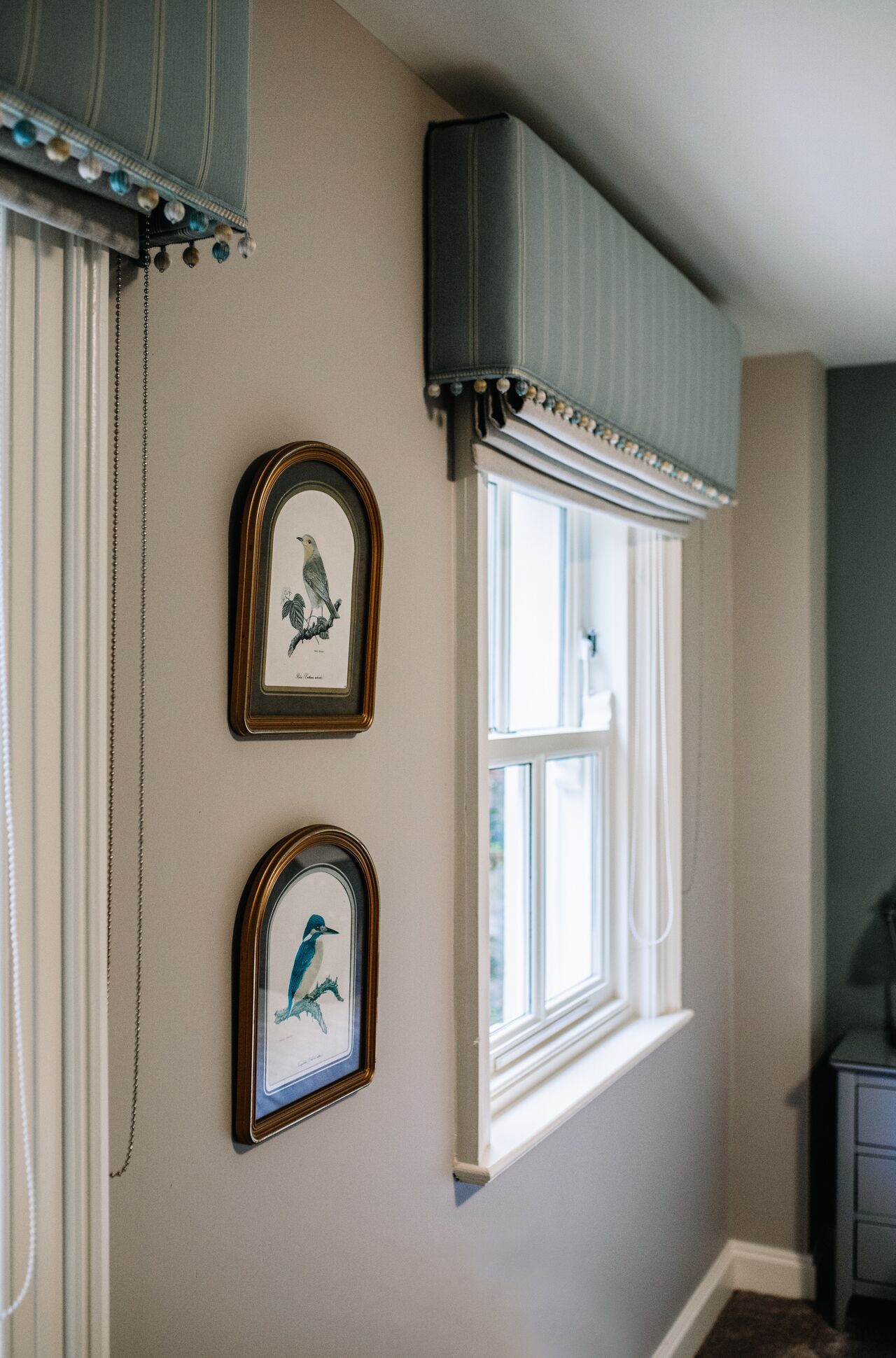 Una habitación con ventana, dos cuadros de pájaros y una cortina.