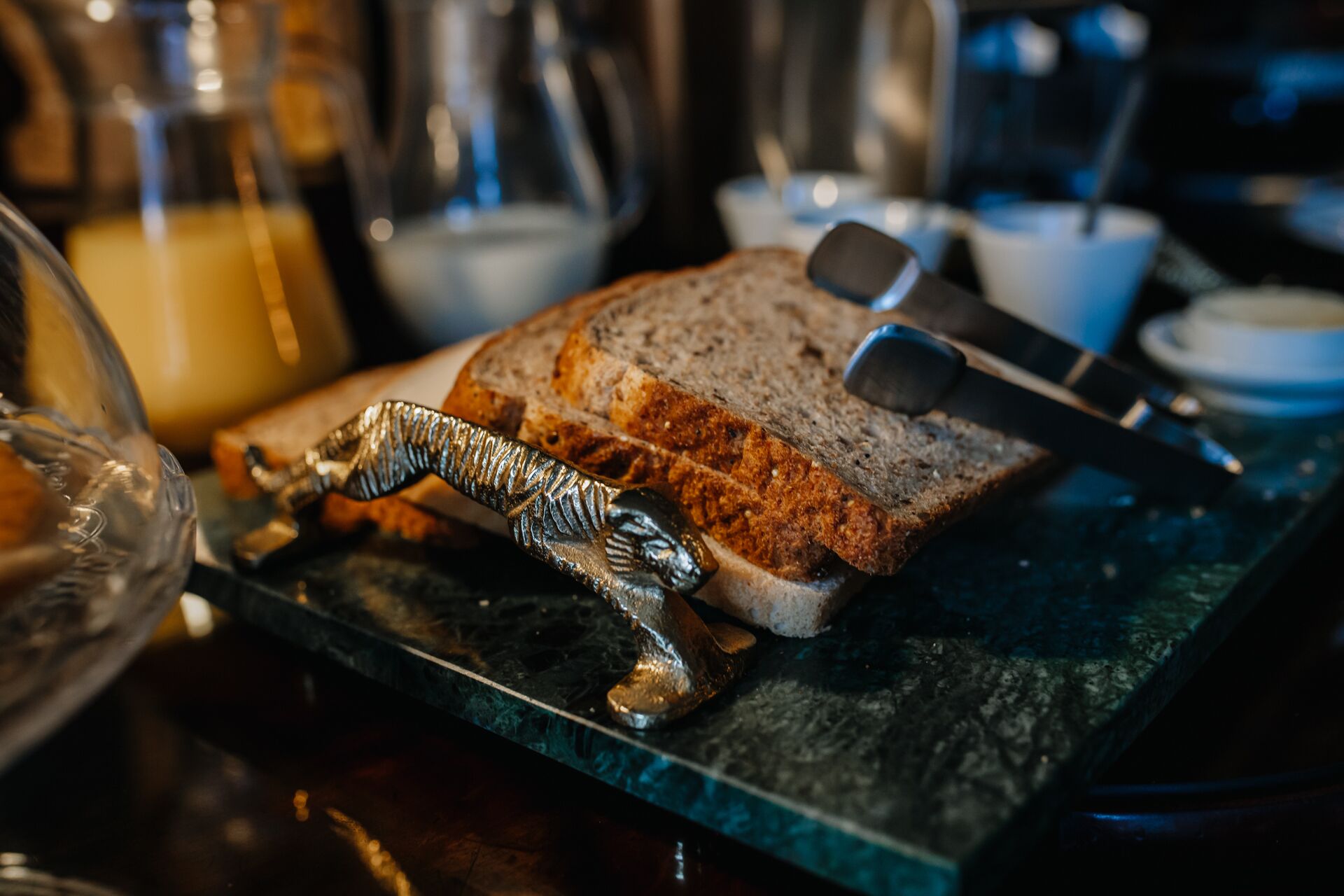 A slice of whole grain bread on a marble countertop with a metal butter knife and a decorative metal spoon.