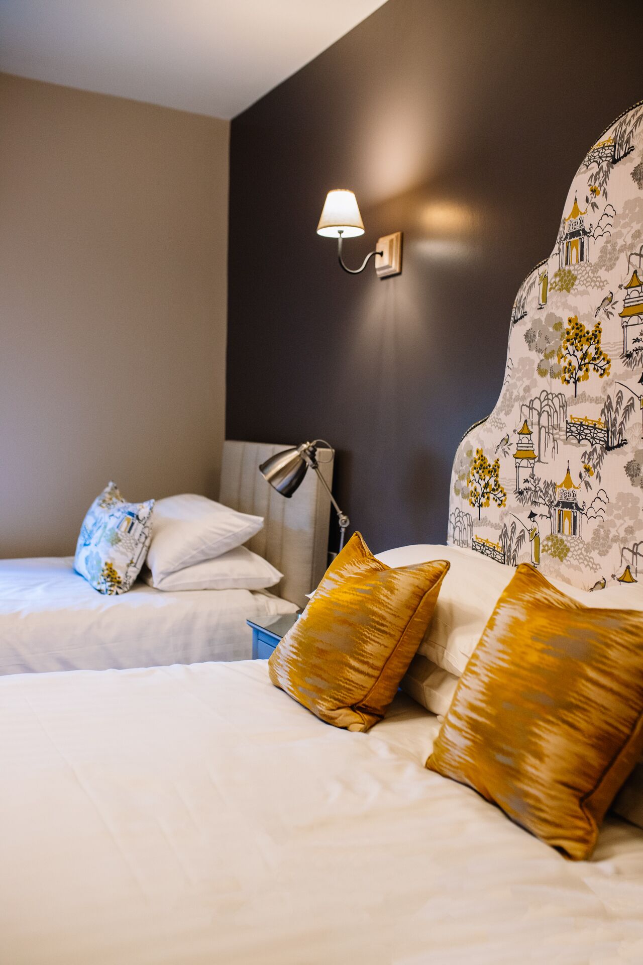 A hotel room with two beds, a nightstand, a lamp, and a wall-mounted light fixture.