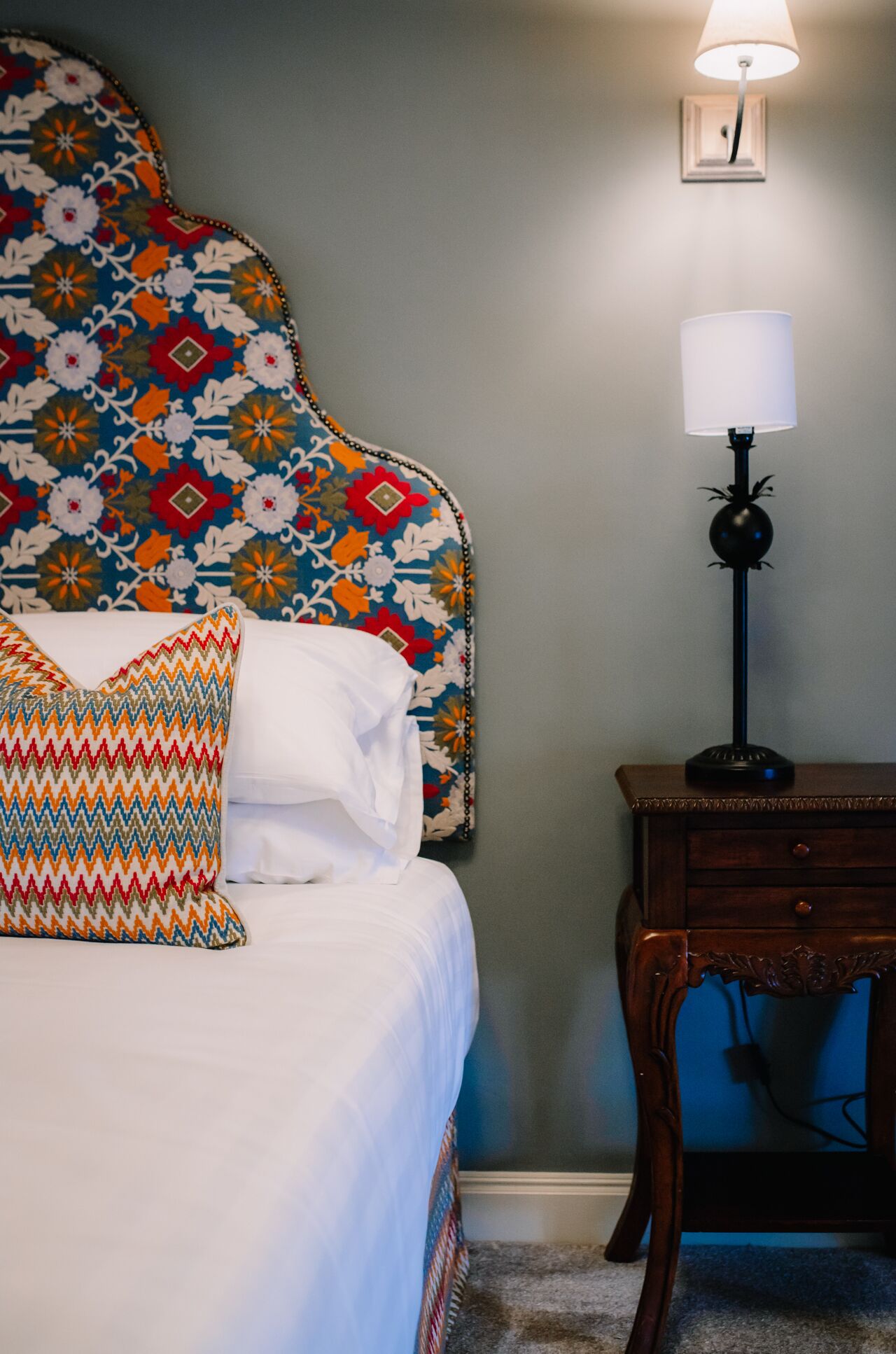 A neatly made bed with white linens and colorful pillows, a patterned headboard, a bedside table with a lamp, and a wall-mounted clock.