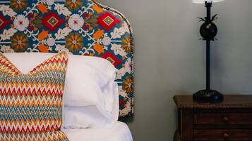 A neatly made bed with white linens and colorful pillows, a patterned headboard, a bedside table with a lamp, and a wall-mounted clock.