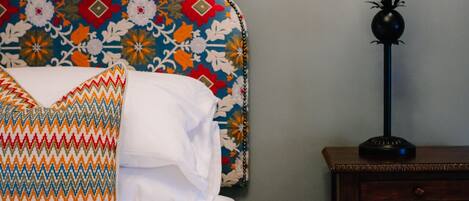 A neatly made bed with white linens and colorful pillows, a patterned headboard, a bedside table with a lamp, and a wall-mounted clock.