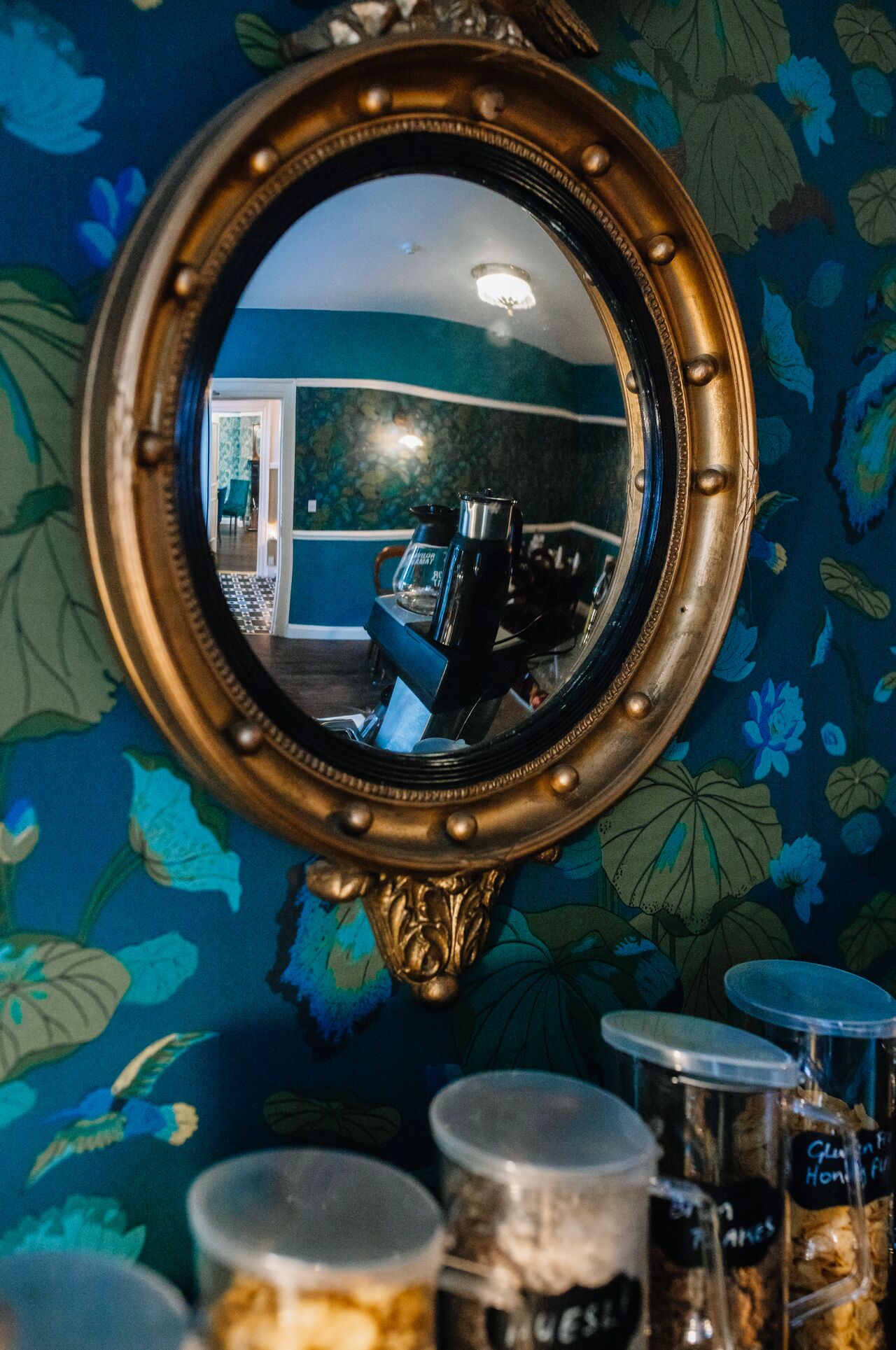 A mirror with a floral wallpaper background and a kitchen area visible in the reflection.