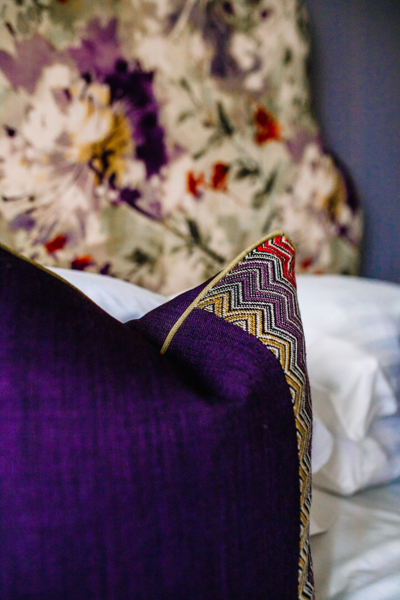 A purple pillow with a patterned edge and a floral-patterned pillow in the background.