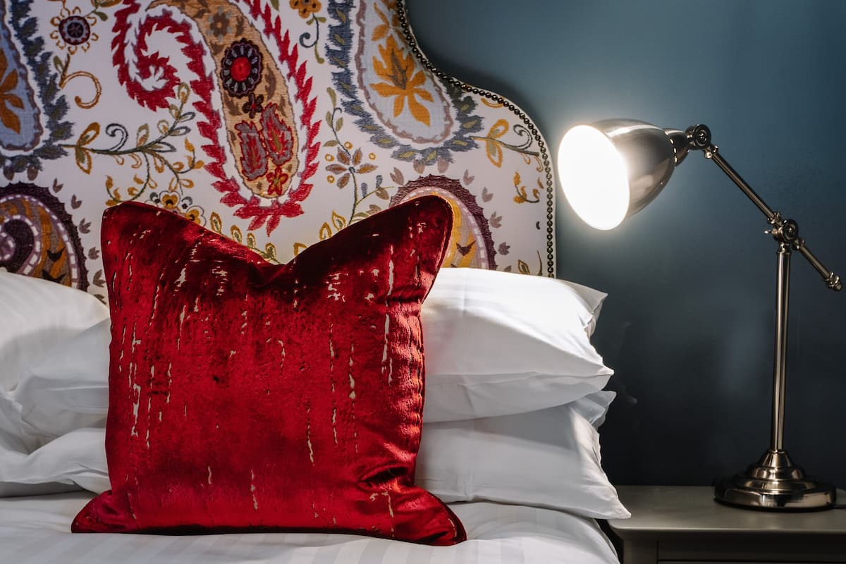 A bed with white linens, a red decorative pillow, and a bedside lamp.