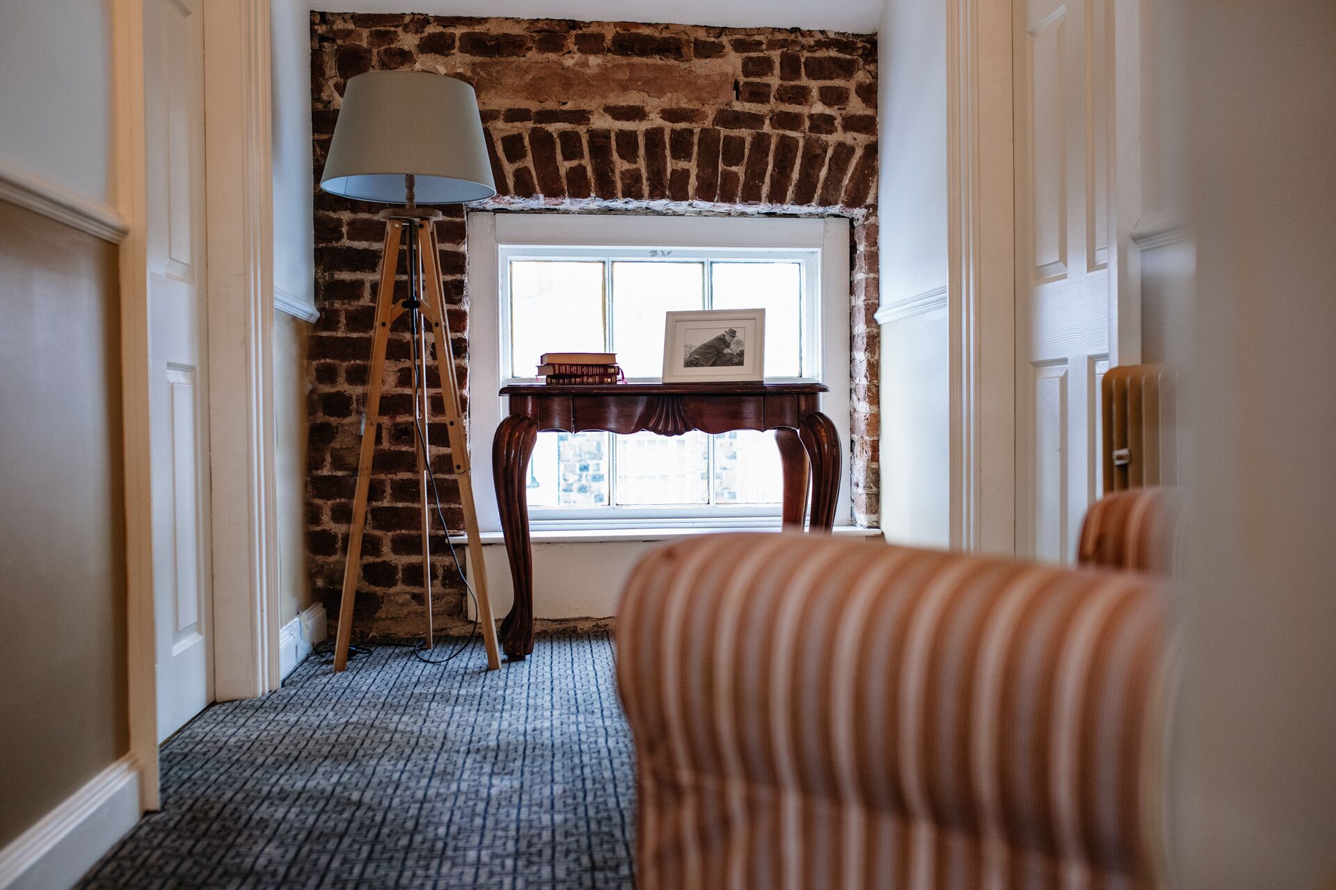 A hallway with a brick wall, a wooden ladder, a table with books and a framed picture, and a striped sofa.