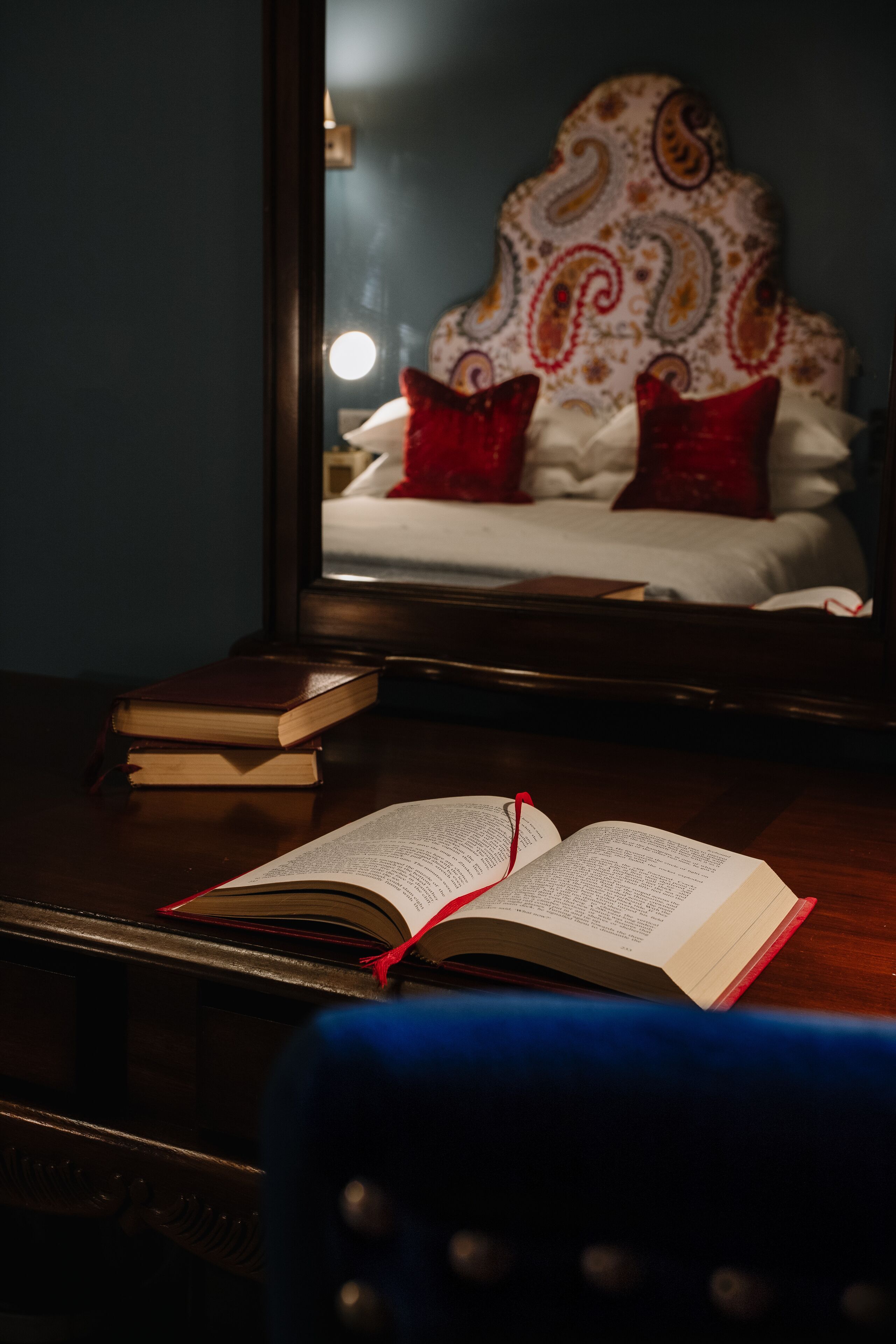 A room with a wooden desk, an open book, and a mirror reflecting a bed with patterned pillows.