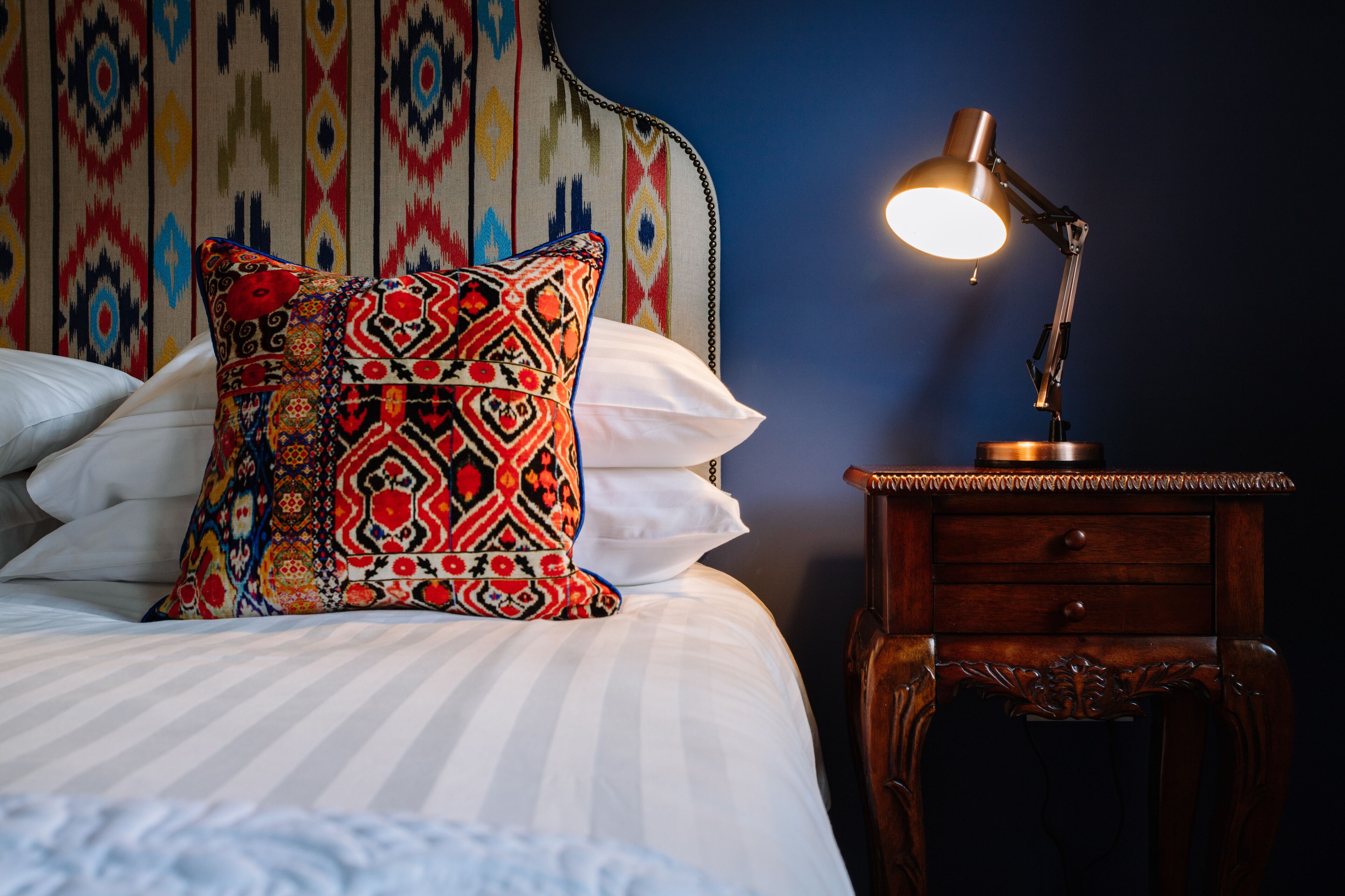 A neatly made bed with a colorful patterned pillow, a bedside table with a lamp, and a headboard with a geometric pattern.
