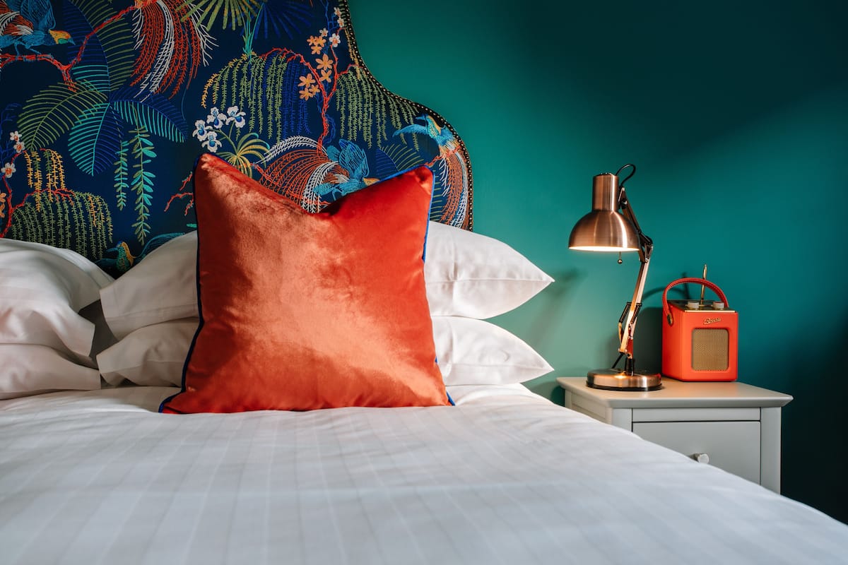 A neatly made bed with white linens and a red pillow, a bedside lamp, and a red radio on a nightstand.