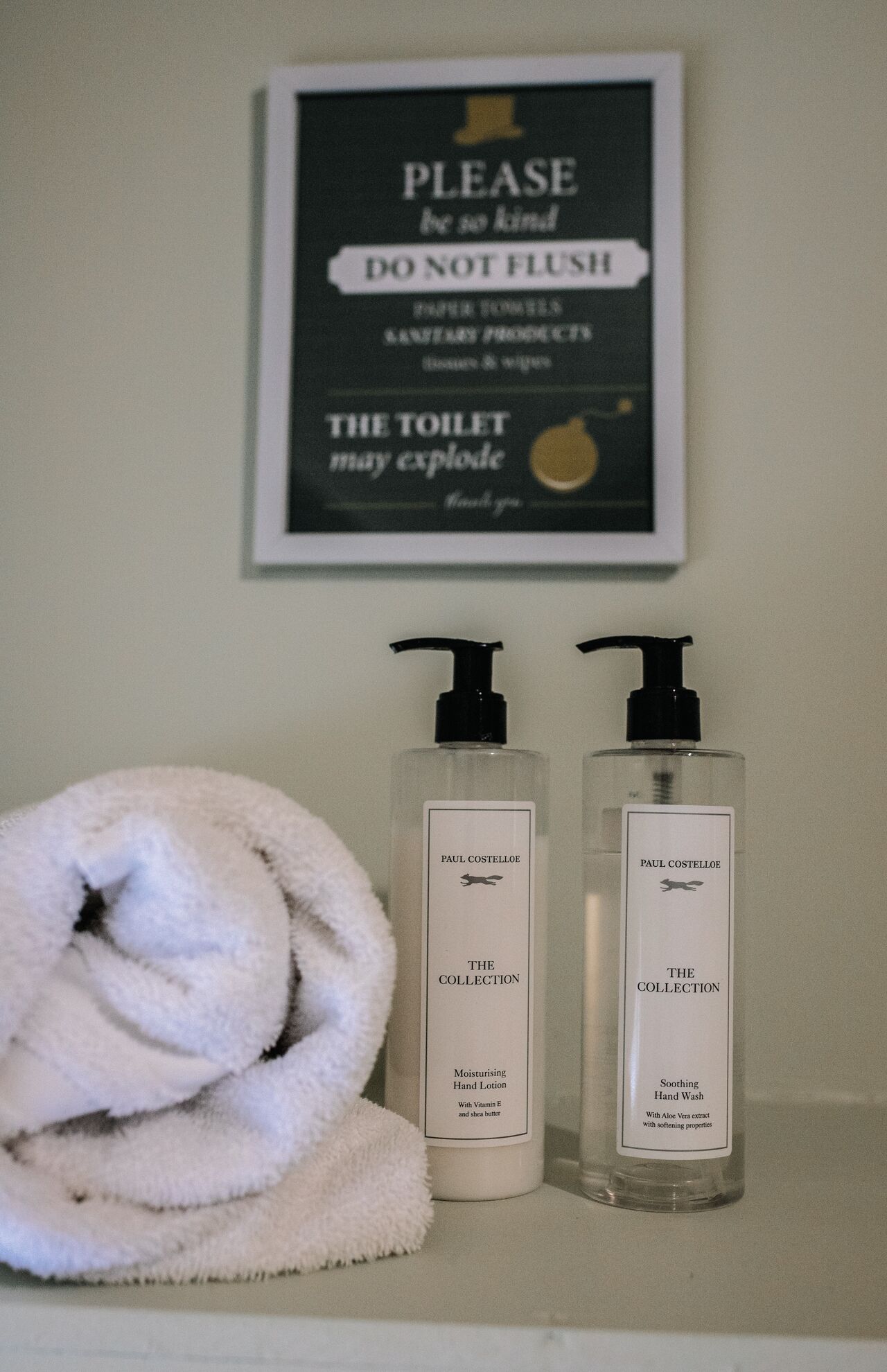 Baño con letrero que indica no tirar toallas de papel, dos productos para el cuidado de manos Paul Costello Collection y una toalla blanca.