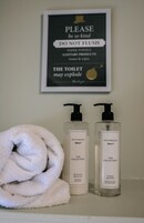 Baño con letrero que indica no tirar toallas de papel, dos productos para el cuidado de manos Paul Costello Collection y una toalla blanca.