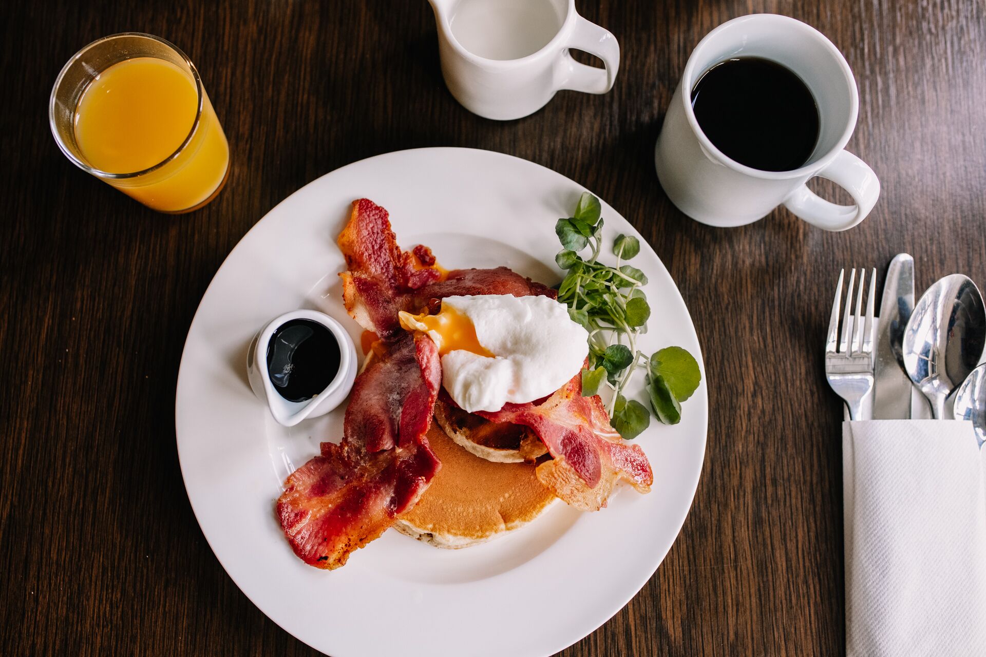 Un plato de desayuno con tocino, huevos y tostadas, acompañado de un vaso de jugo de naranja y dos tazas de café sobre una mesa de madera.