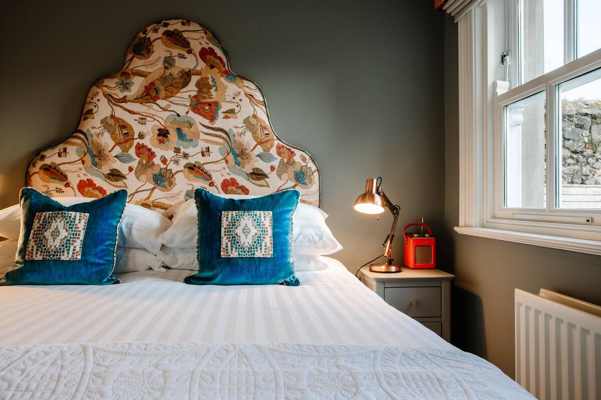 A neatly made bed with a floral headboard, two blue pillows, a bedside lamp, and a window with a view of a stone wall.