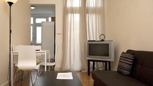 A living room with a black coffee table, a brown sofa, a TV on a stand, and a white chair. - Tooting Place Apartments (Mitcham)