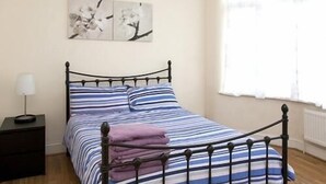 A bed with a metal frame, a striped bedspread, and a pillow. There is a nightstand with a lamp beside the bed. A window with curtains is visible in the background. - Tooting Place Apartments (Mitcham)