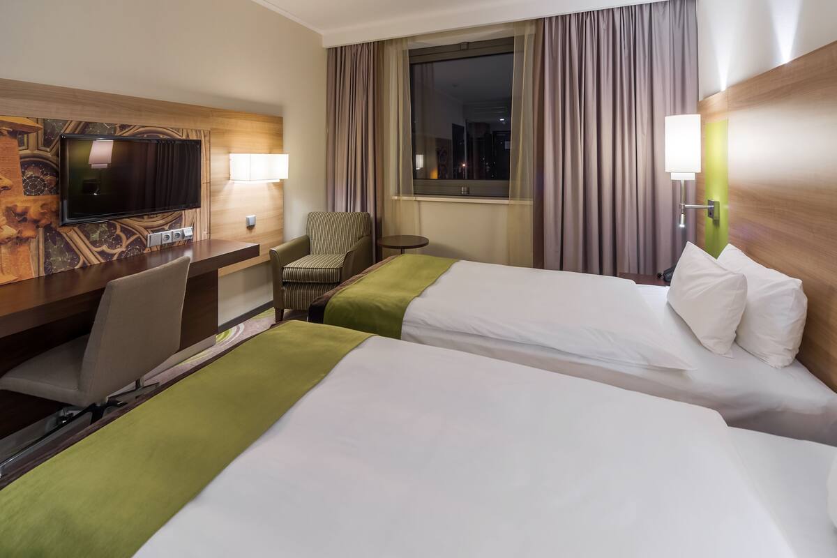 A hotel room with two beds, a desk with a television, a chair, a small table, and a window with curtains.