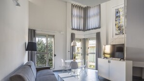 Una sala de estar moderna con un sofá gris, una mesita blanca pequeña y un televisor sobre un mueble blanco. Hay dos ventanas con cortinas, una zona de comedor con mesa y sillas, y un cuadro en la pared.