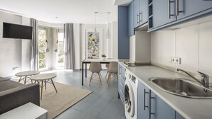 Una cocina moderna con armarios azules, un lavamanos de acero inoxidable y una lavadora integrada. Hay una zona de comedor con una mesa y sillas, y un televisor montado en la pared.