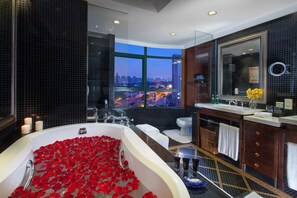 Un baño moderno con una bañera grande llena de pétalos de rosa, un ventanal con vistas a la ciudad y un tocador con espejo y lavabo.