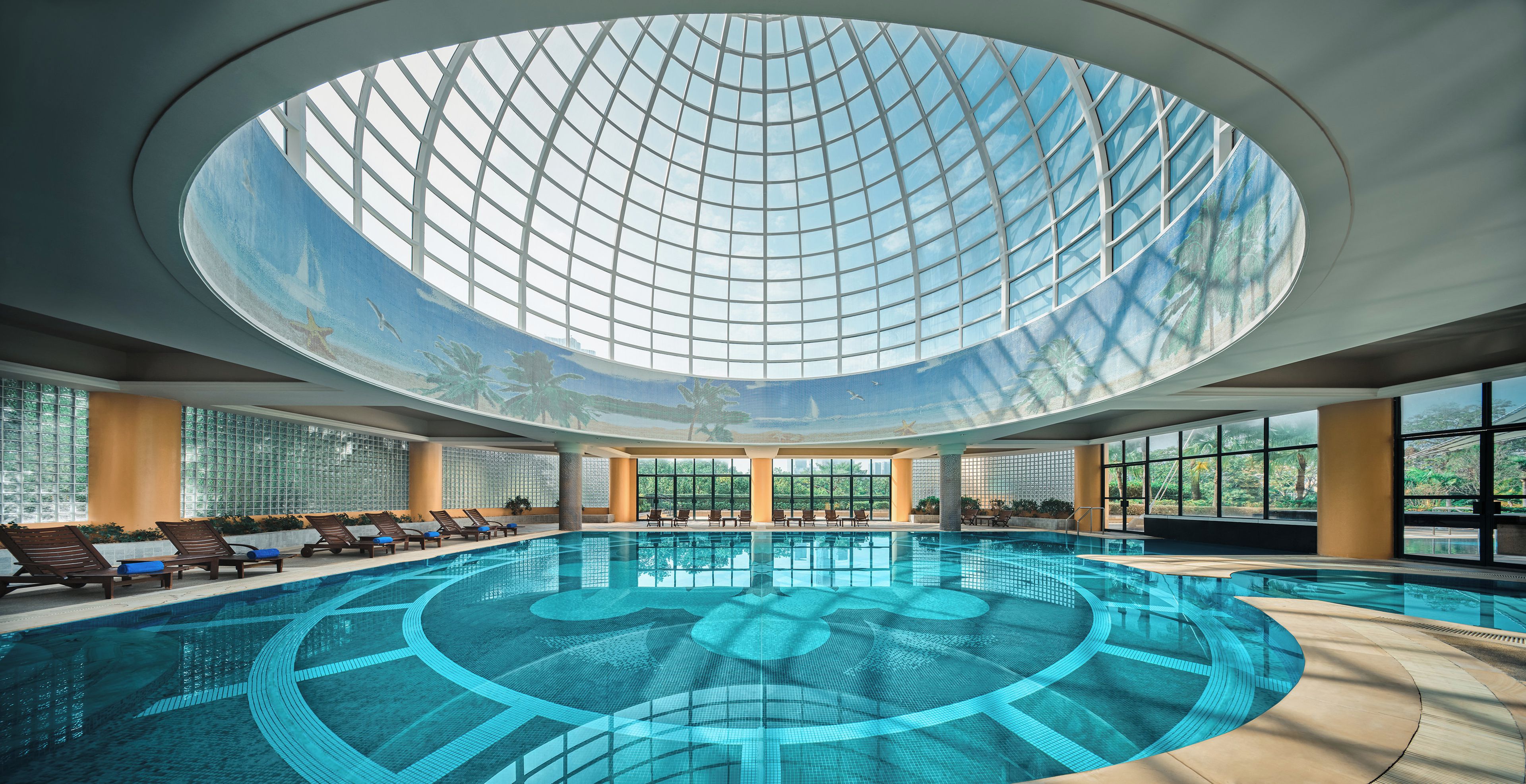 A large indoor swimming pool with a glass ceiling, surrounded by lounge chairs and a view of greenery outside.