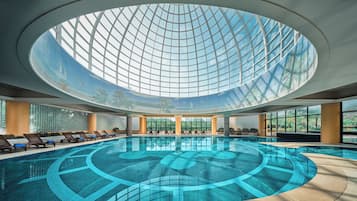 A large indoor swimming pool with a glass ceiling, surrounded by lounge chairs and a view of greenery outside.