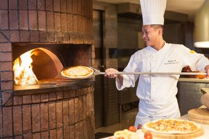 A chef in a white uniform with a chef's hat, using a long-handled tool to retrieve a pizza from a brick oven.