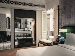 Una habitación de hotel moderna con una cama grande, una mesita de noche con lámpara, un baño con espejo y lavabo, y vistas a la ciudad desde la ventana.