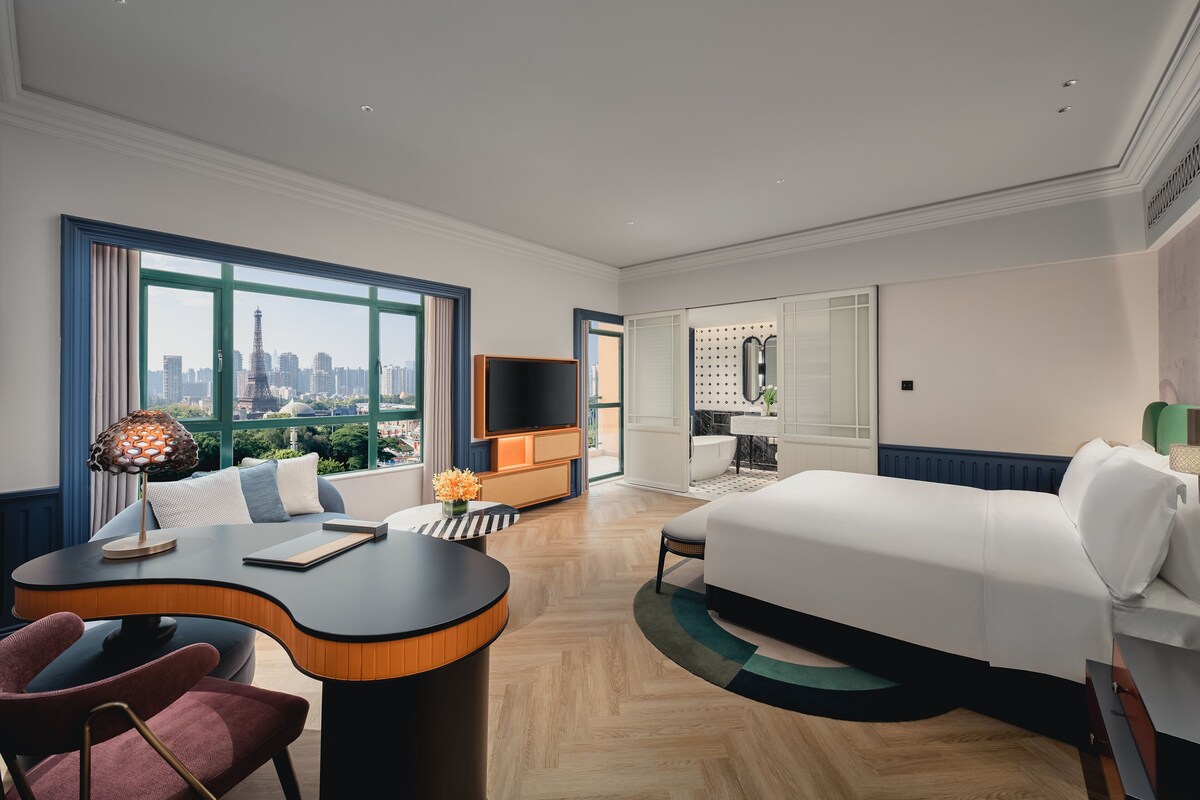 A modern hotel room with a large bed, a desk with a lamp, a view of the city, and a bathroom with a bathtub.