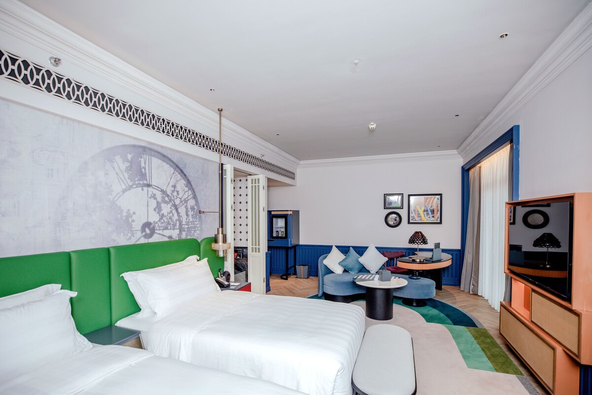 A hotel room with a large bed, a green headboard, a seating area with a small table, a television, and artwork on the walls.
