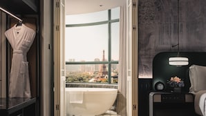 Un baño moderno con una bañera grande, una bata colgada en un perchero y vistas de un paisaje urbano a través de un ventanal.