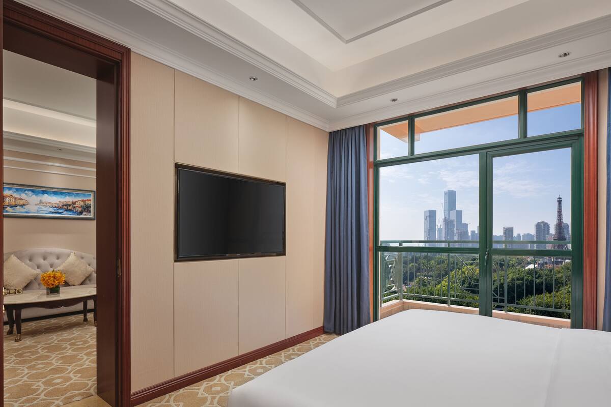 Ein Hotelzimmer mit einem großen Bett, einem wandmontierten Flachbildfernseher, einer Couch mit einer Blumenvase und Blick auf die Stadt durch große Fenster.