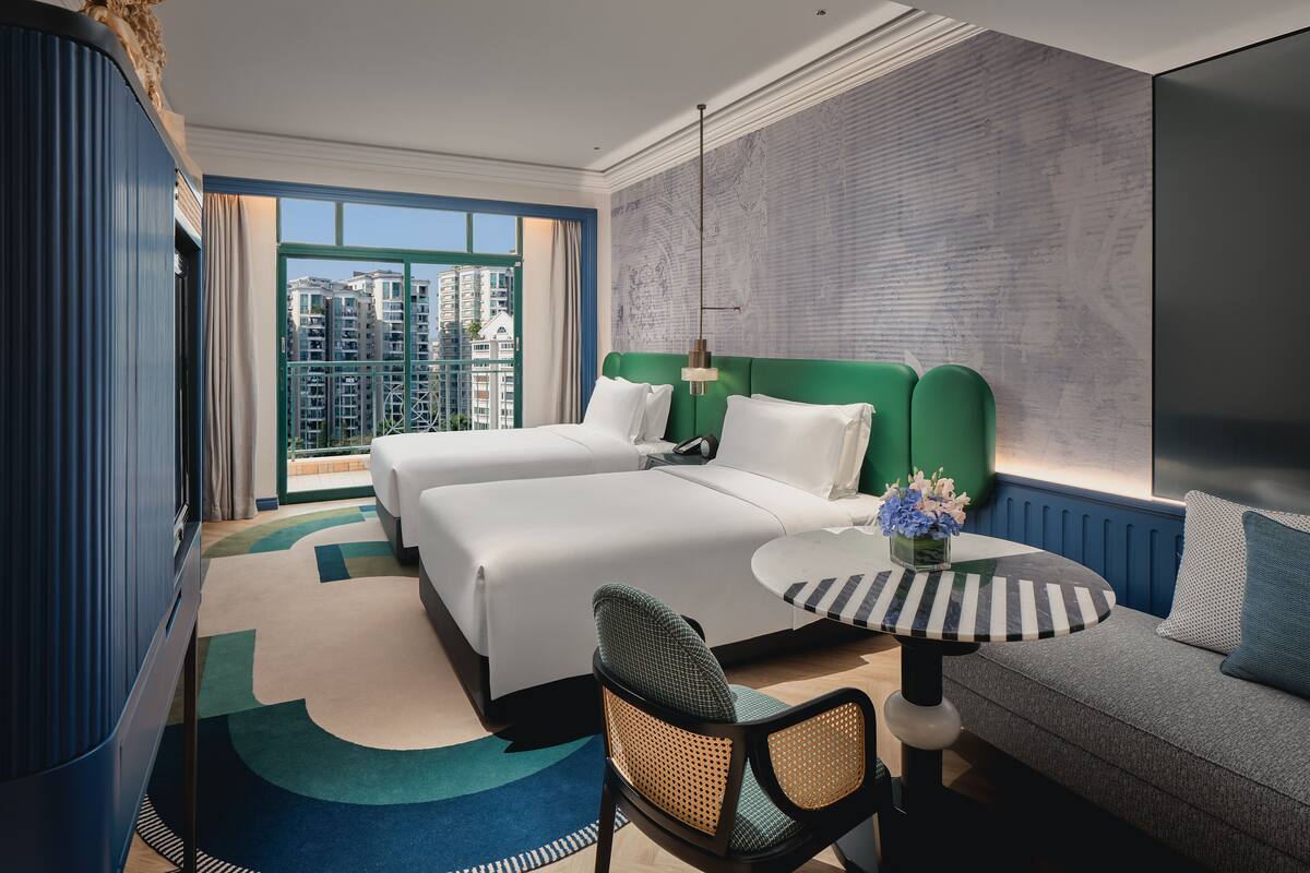 Ein Hotelzimmer mit zwei Betten, einem Esstisch, einer Couch und einem großen Fenster mit Blick auf die Stadt.