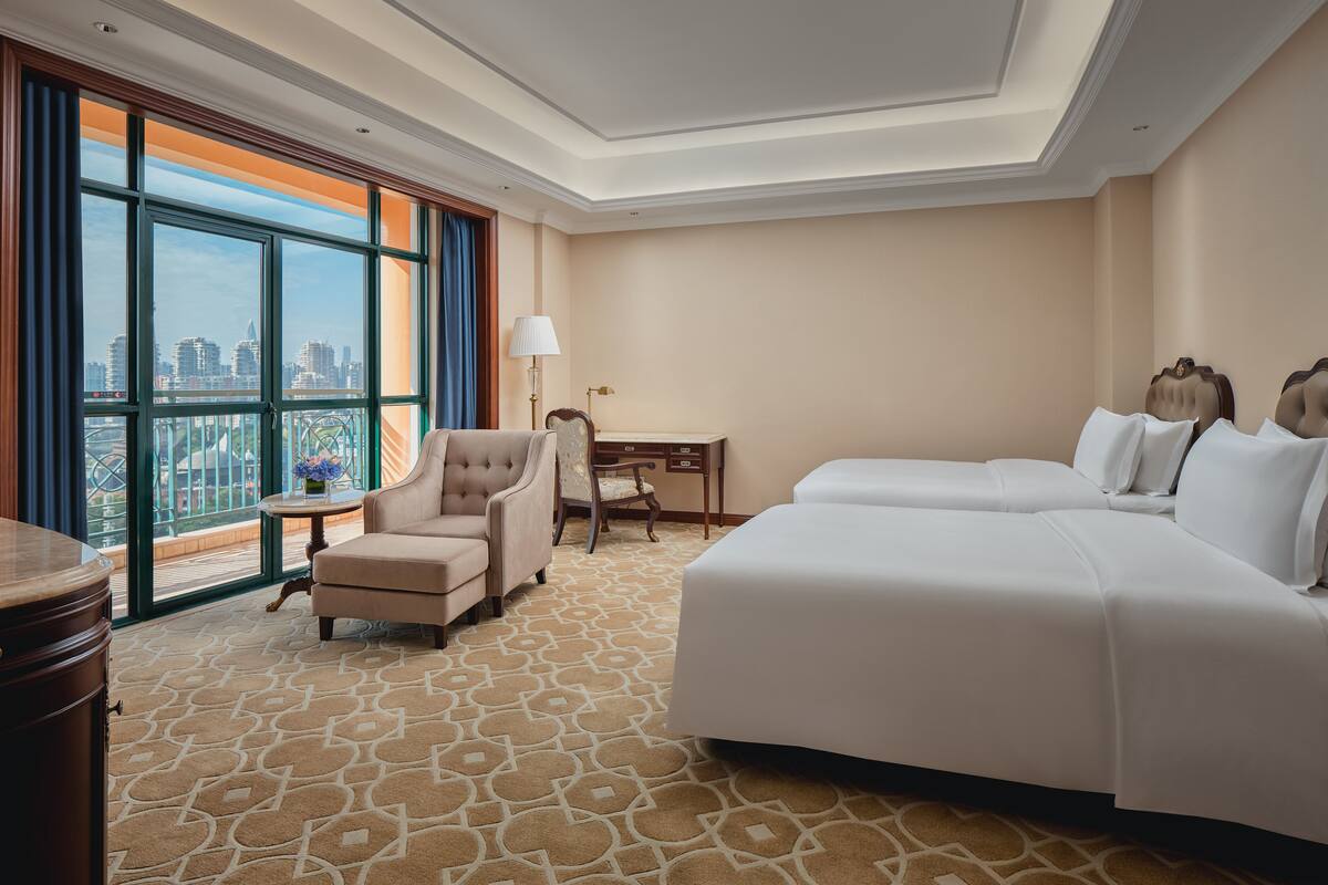 Ein Hotelzimmer mit großem Fenster, Schreibtisch, zwei Betten und einem Sessel.