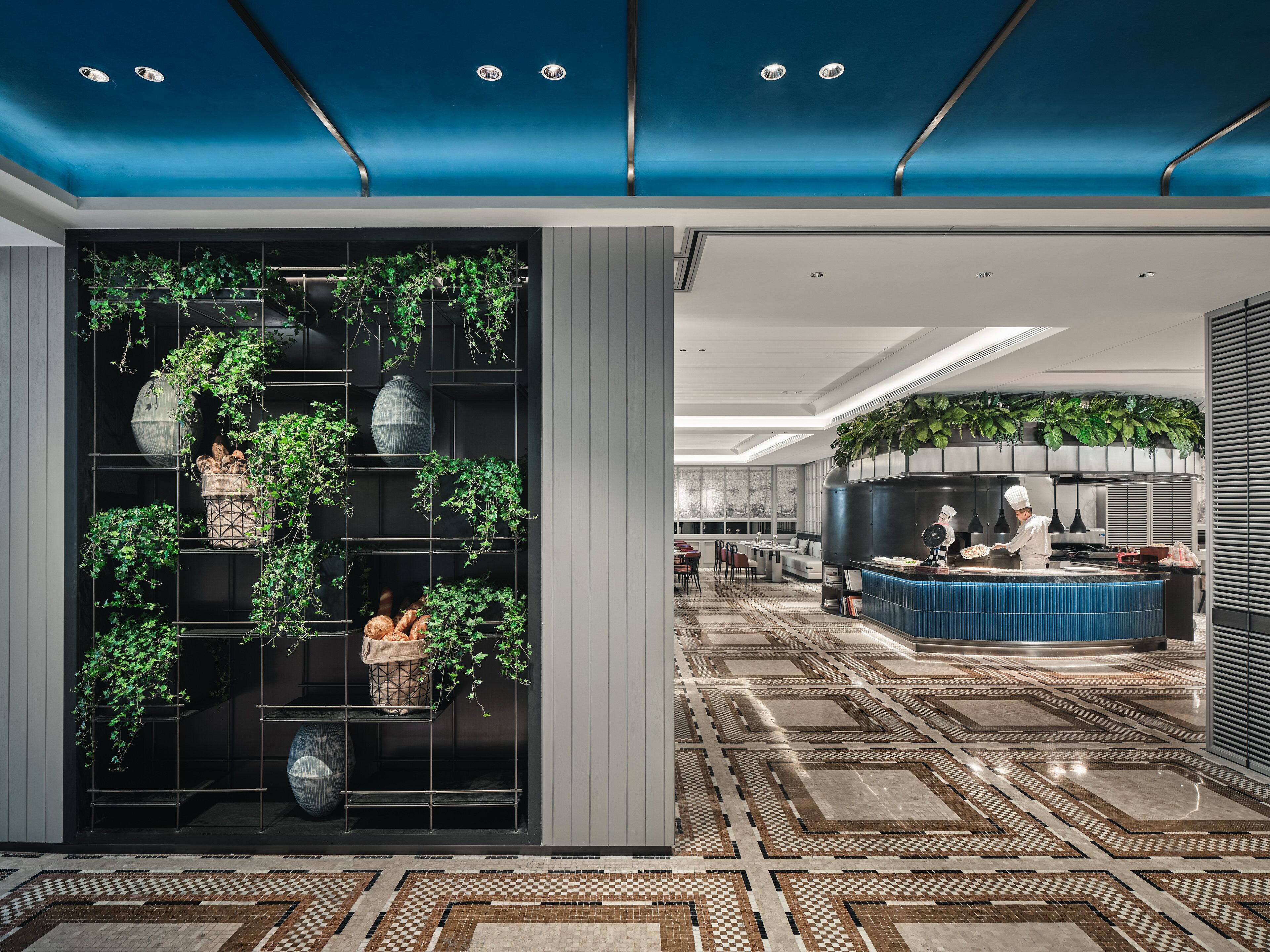 Um hall de hotel moderno com um grande arranjo de plantas, uma área de cozinha e um chão estampado.