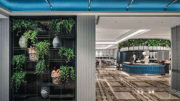 Um hall de hotel moderno com um grande arranjo de plantas, uma área de cozinha e um chão estampado.