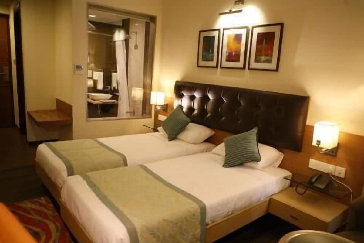 Quarto de hotel com duas camas, um espelho, mesa de cabeceira com abajur e janela com vista para um banheiro.
