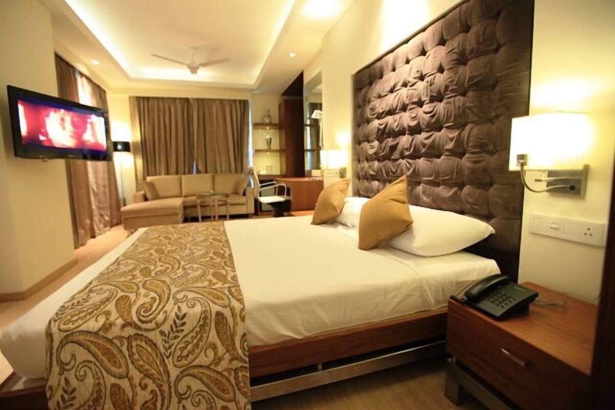 Quarto de hotel com cama, mesa de cabeceira, televisão e área de estar com sofá e cadeiras.