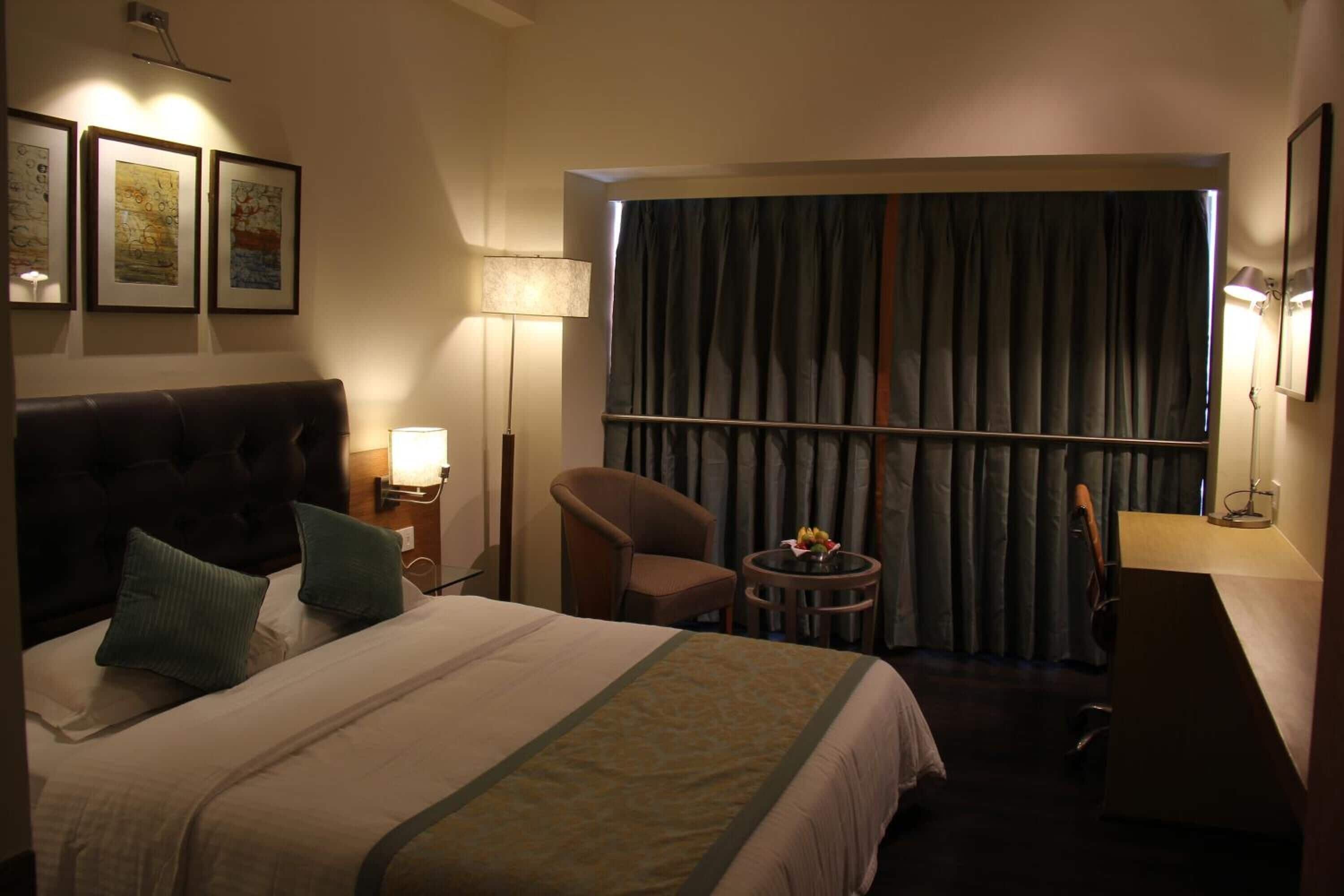 Habitación de hotel con una cama grande, una silla, una mesita con un florero y vistas al exterior a través de una ventana con cortinas.