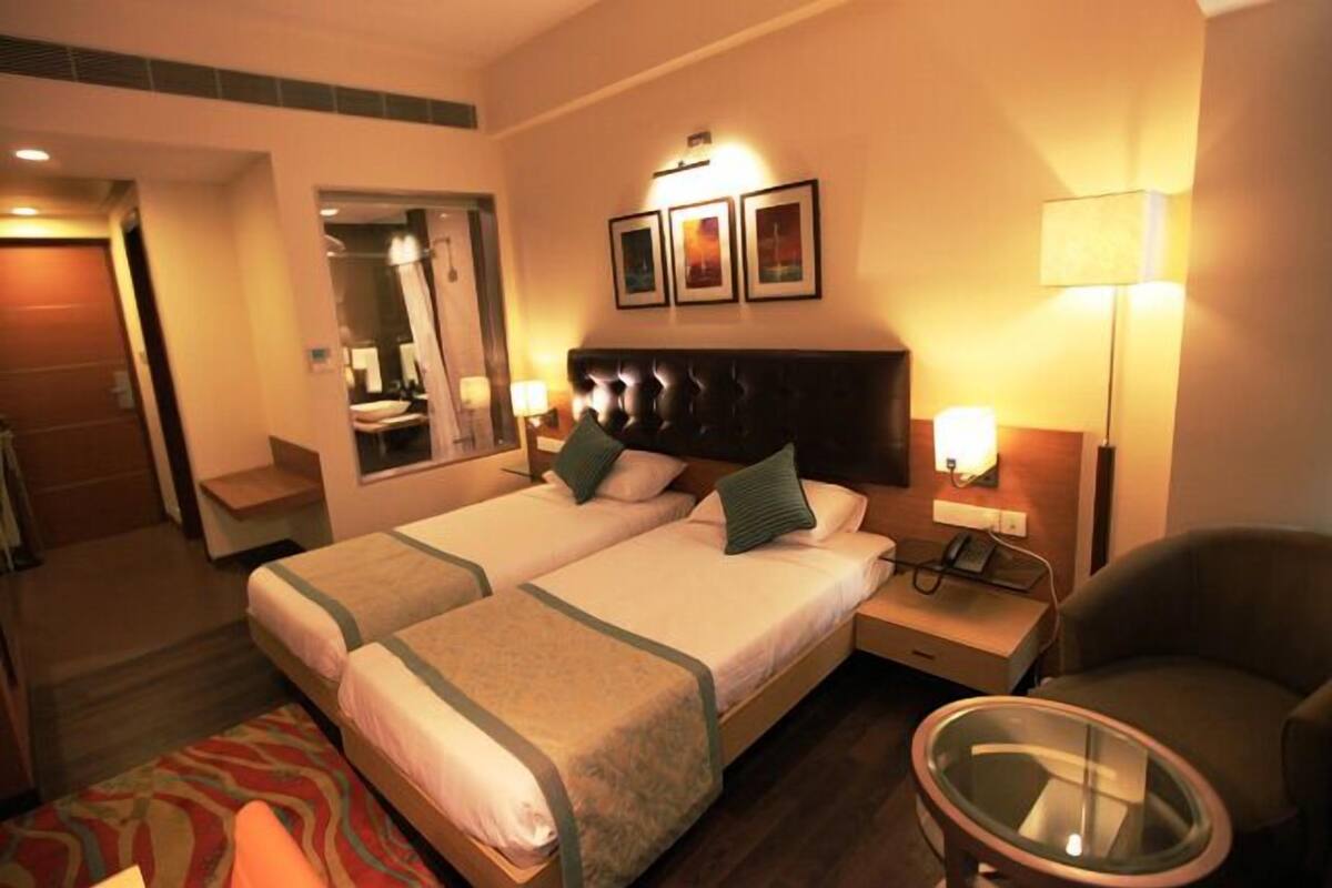 Quarto de hotel com duas camas, um espaço de estar com mesa de vidro, um espelho e uma luminária na parede.