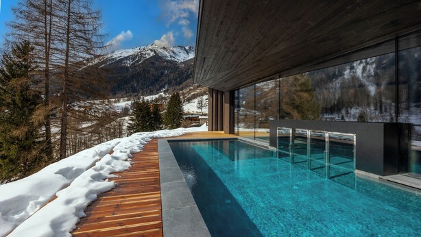 Ein moderner Außenpool mit Holzterrasse und Glaswänden, umgeben von schneebedeckten Bergen und Bäumen.
