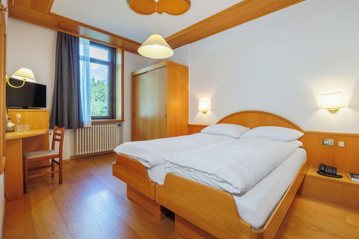 Een slaapkamer met houten frame, een bed, bedlampjes, een bureau met lamp, een stoel en een raam met gordijnen.
