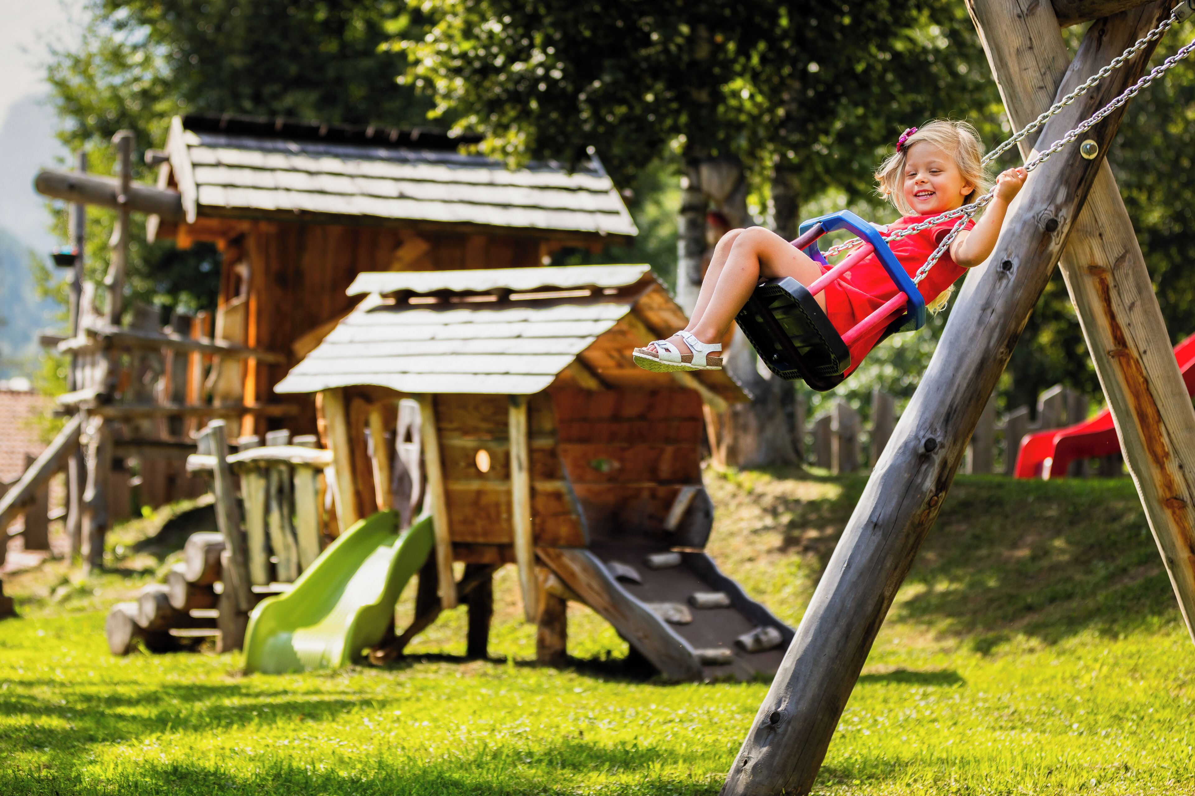 A child on a swing in a playground with a slide and wooden play structures.