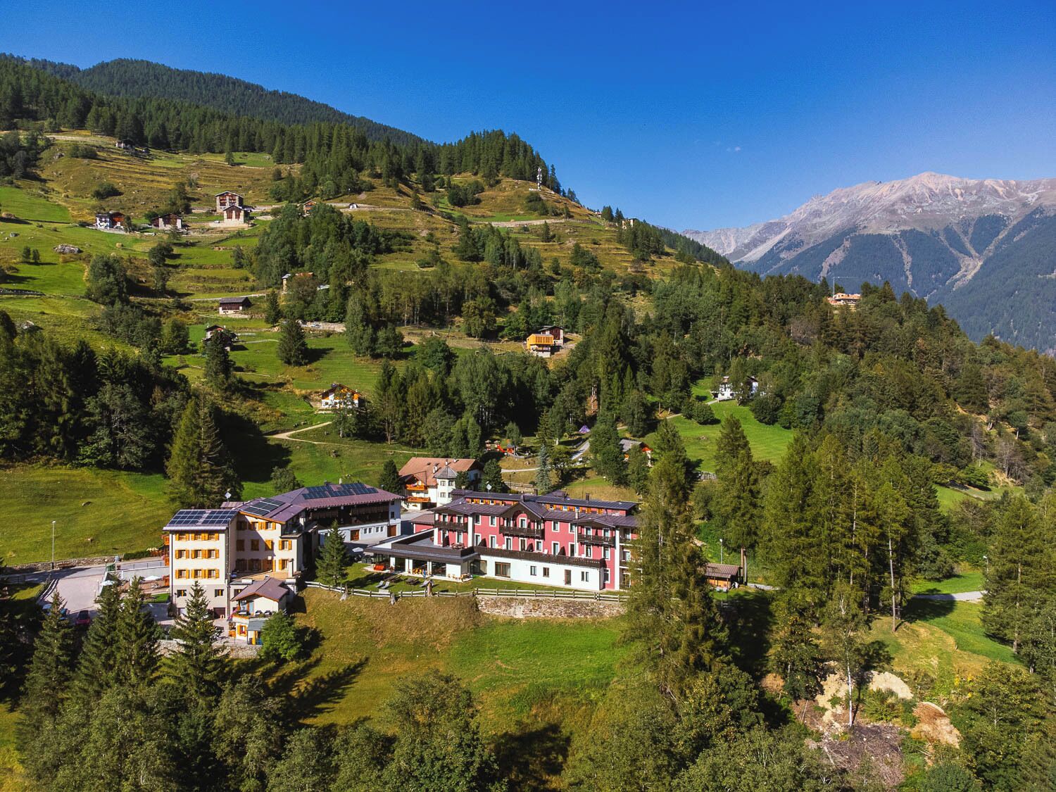 A hillside village with traditional buildings, a prominent multi-story hotel with a red facade, and a clear view of the surrounding mountains.