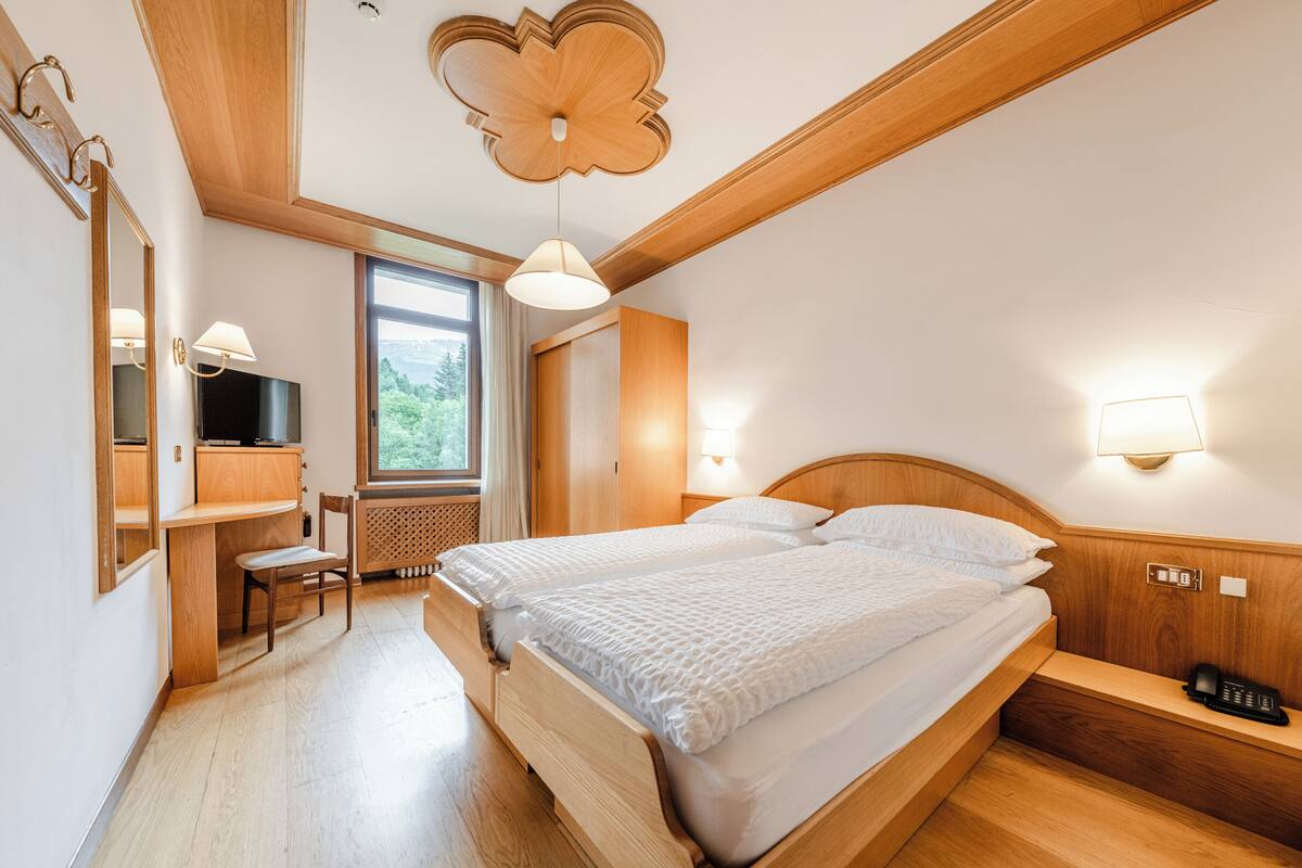 Een slaapkamer met een houten bed, een bureau en een stoel. Een raam biedt uitzicht op groen buiten.