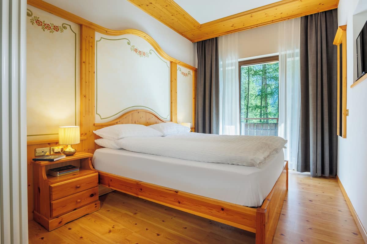 Een houten bed met hoofdeinde, een nachtkastje met een lamp en een raam met gordijnen.