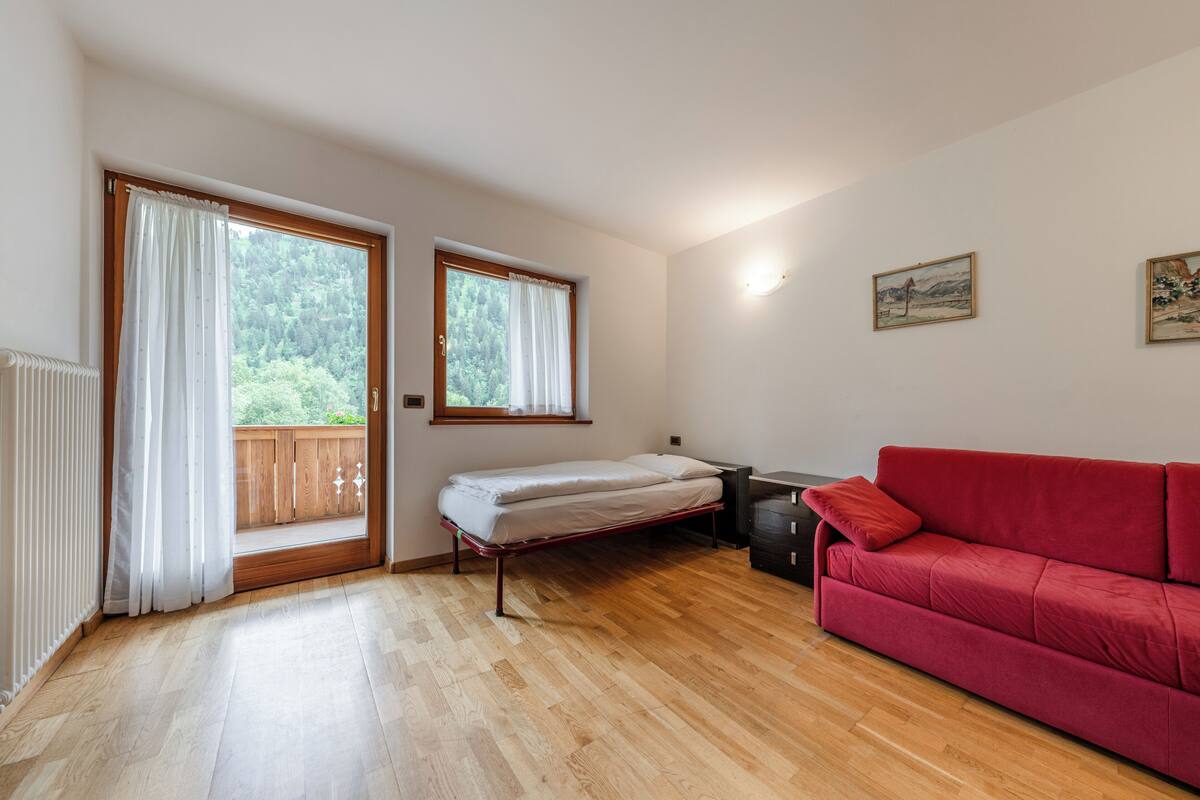 Een kamer met een houten vloer, een rode bank, een bed met witte lakens en twee ramen met gordijnen.
